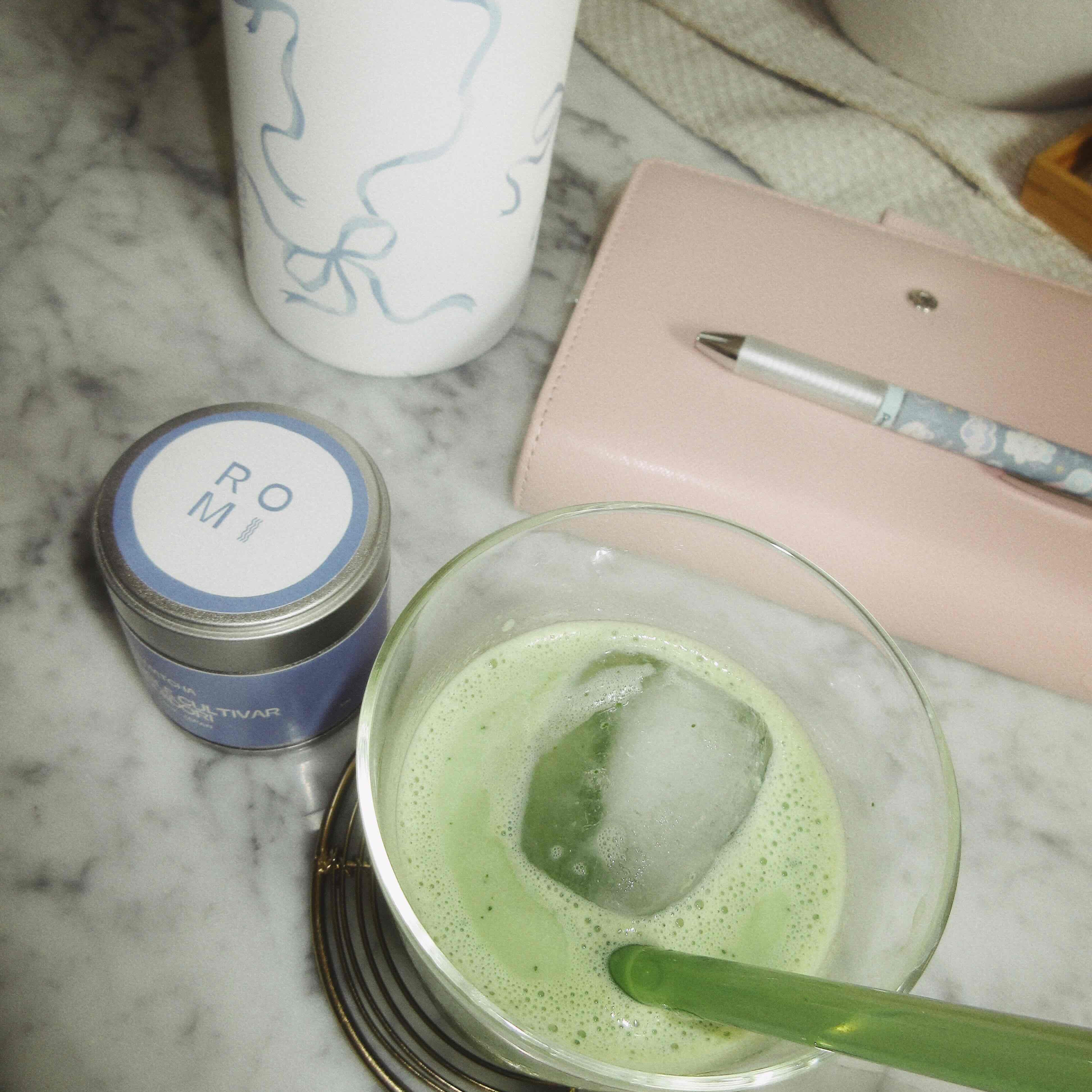 matcha tin powder with matcha latte in a glass with water bottle and notebook