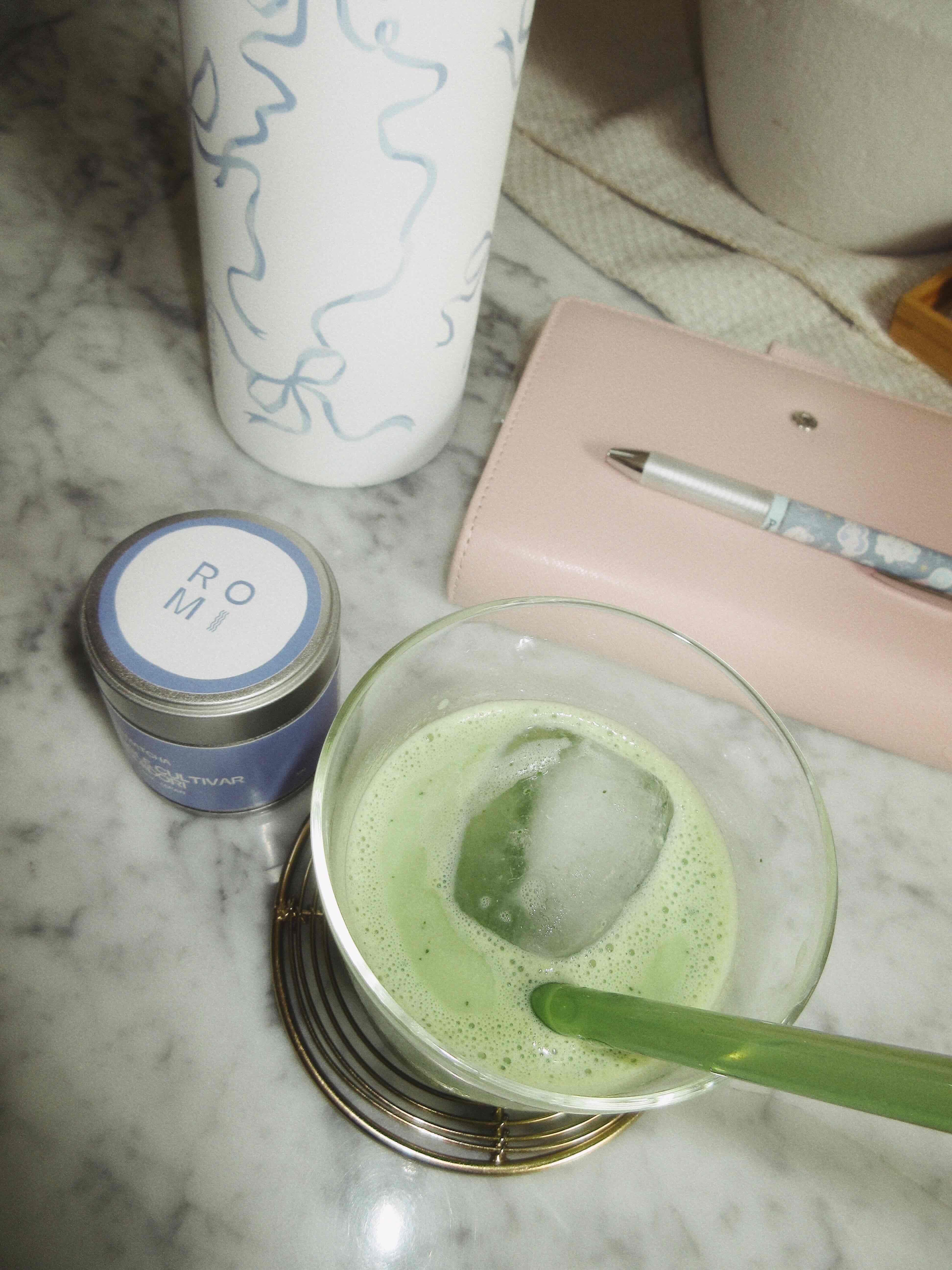 matcha tin powder with matcha latte in a glass with water bottle and notebook