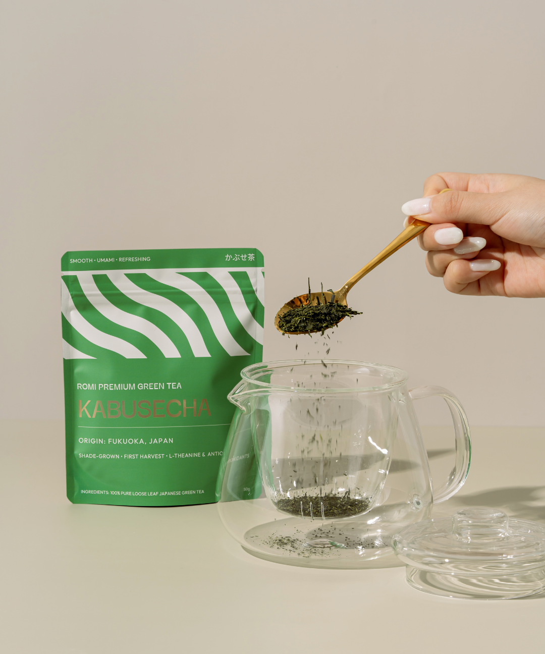 Person adding green tea leaves to a glass teapot with a green tea package labeled 'Kabusecha' in the background.