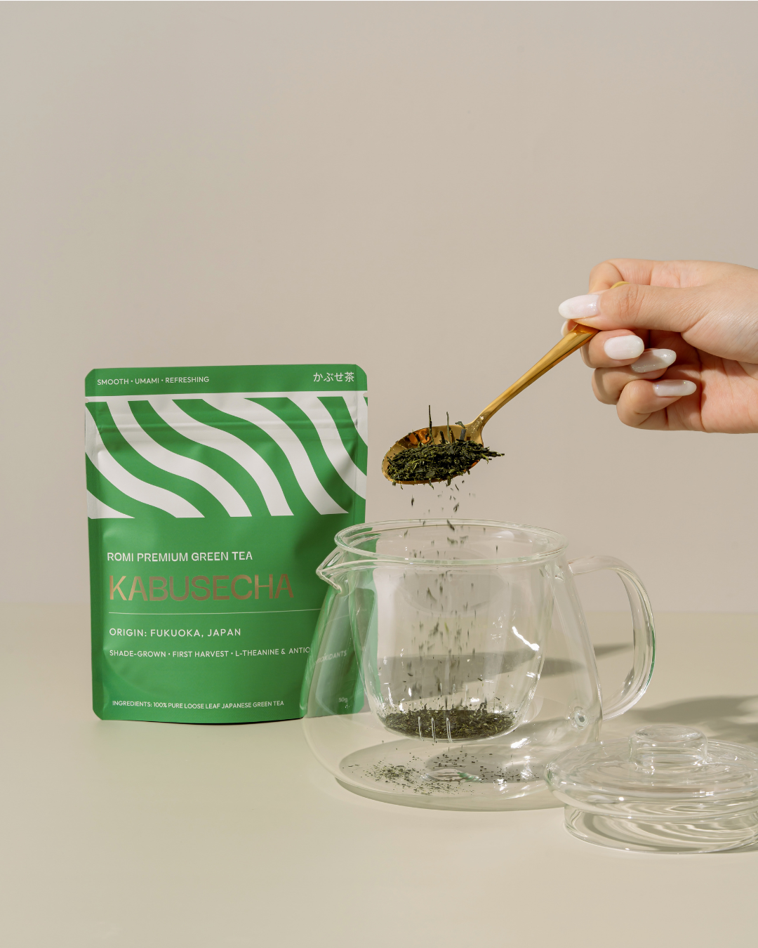 Person adding green tea leaves to a glass teapot with a green tea package labeled 'Kabusecha' in the background.
