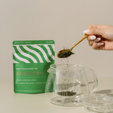 Person adding green tea leaves to a glass teapot with a green tea package labeled 'Kabusecha' in the background.