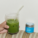 Green smoothie in a glass with a straw, next to a container of matcha powder on a striped surface.