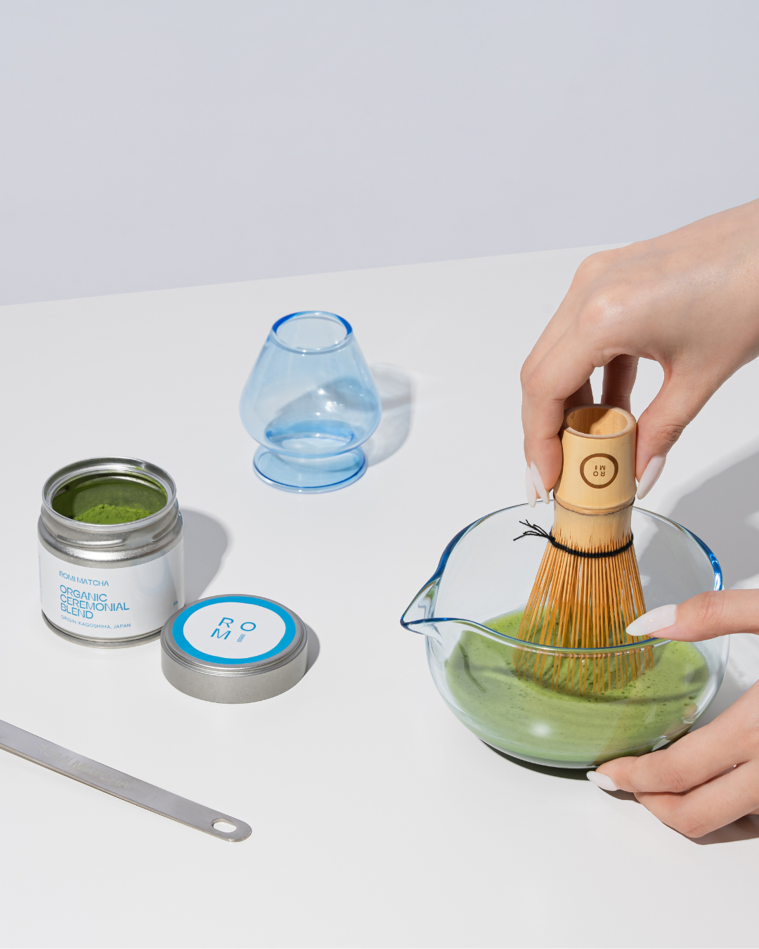 Person using a wooden whisk to mix matcha in a glass bowl with containers and a blue cup in the background.