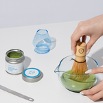 Person using a wooden whisk to mix matcha in a glass bowl with containers and a blue cup in the background.
