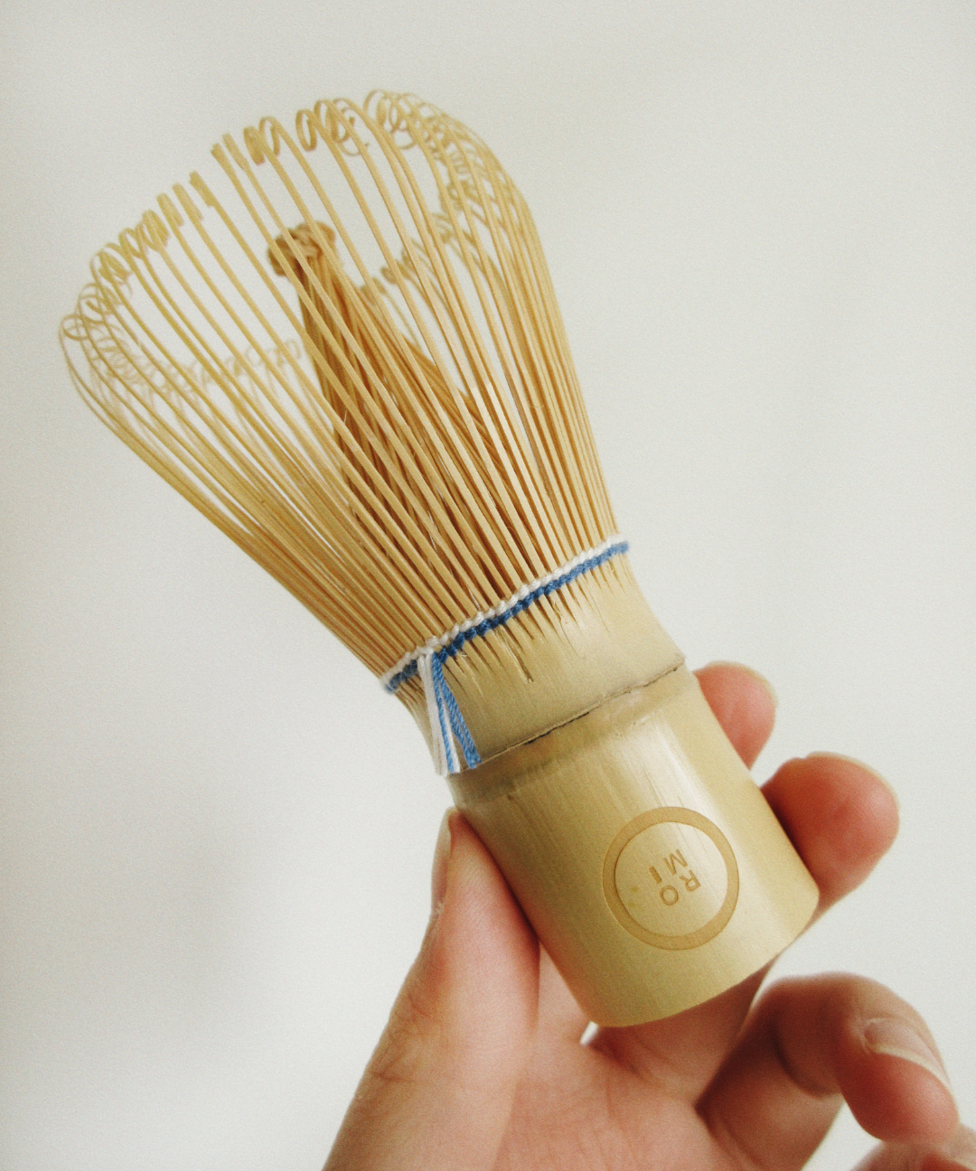 Hand holding a wooden tea whisk against a plain background