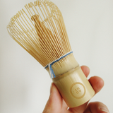 Hand holding a wooden tea whisk against a plain background