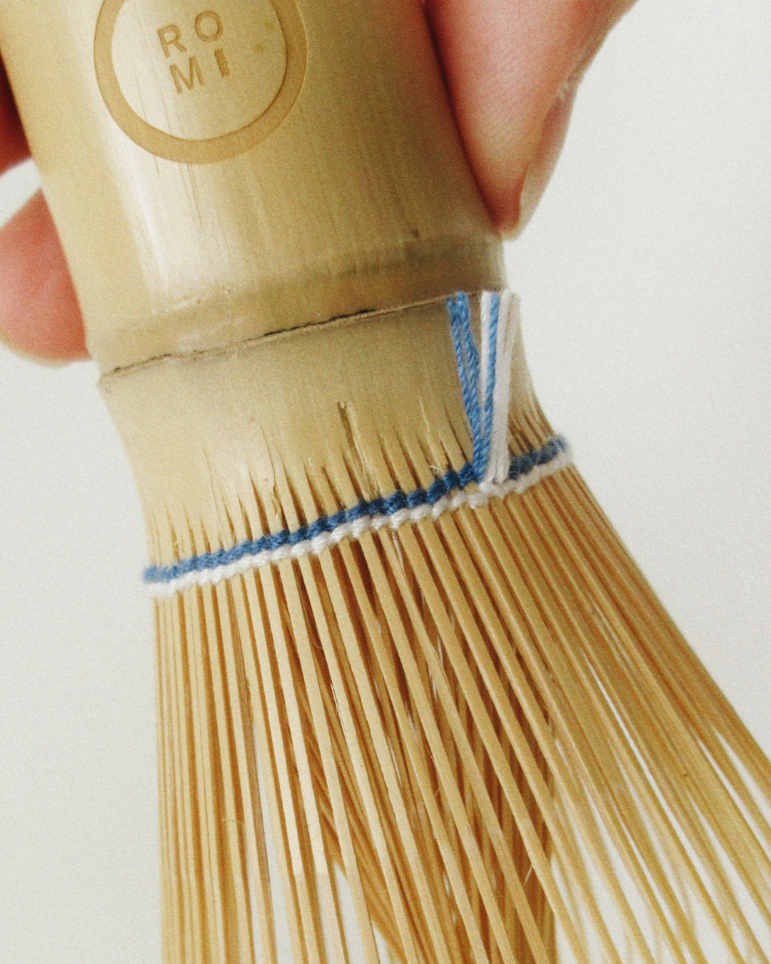 Wooden whisk with blue string, held by a hand against a white background