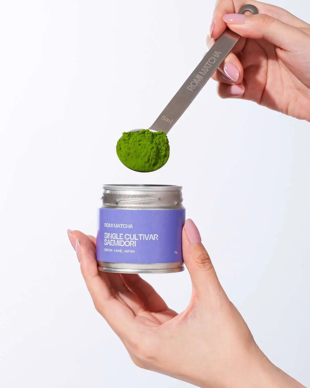 Person holding a container of matcha powder with a spoonful above it on a white background