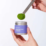 Person holding a container of matcha powder with a spoonful above it on a white background