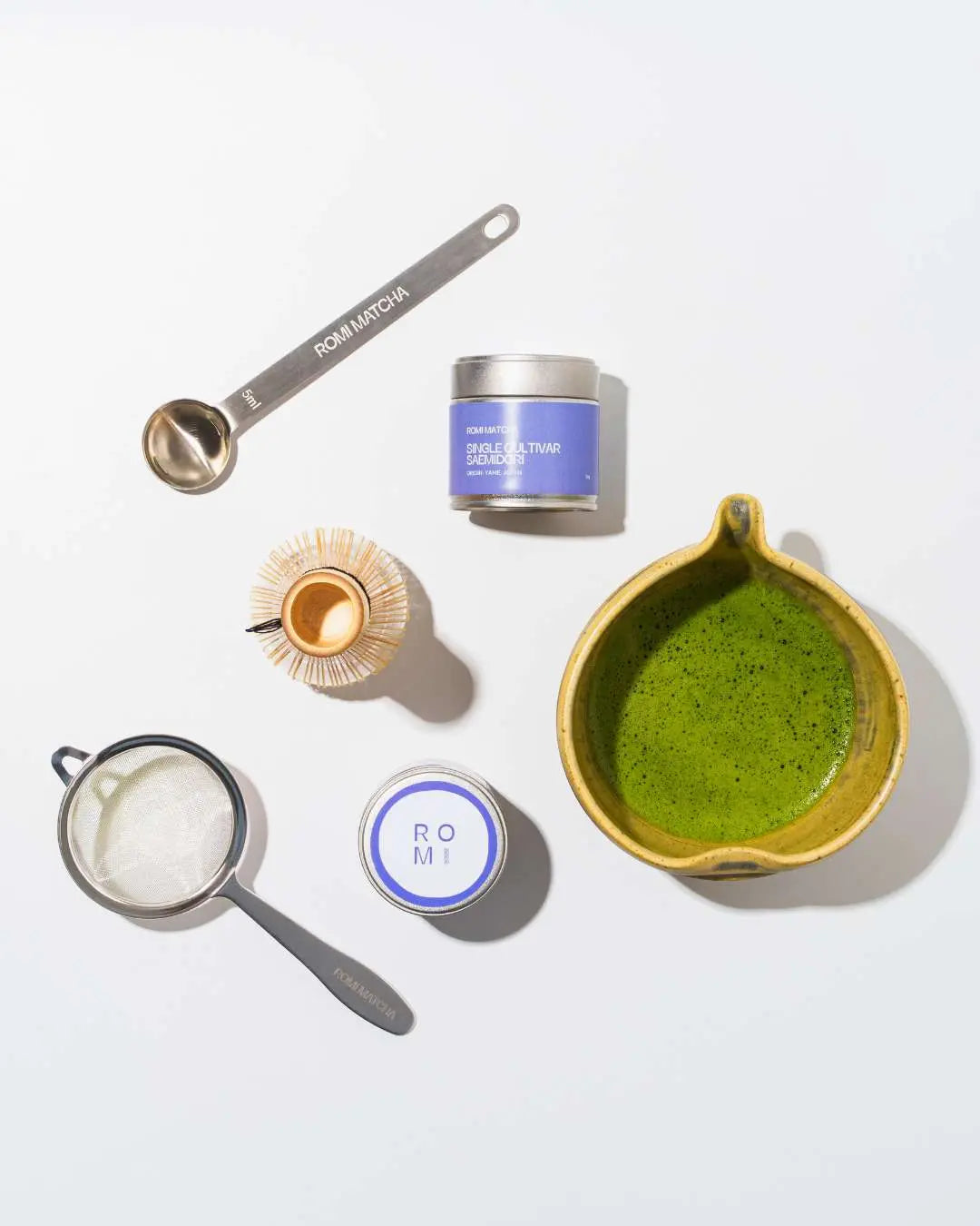 Tea-making tools including a bowl with matcha green tea, a spoon, a whisk, and containers on a white background.