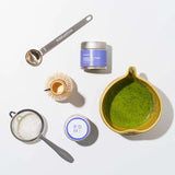 Tea-making tools including a bowl with matcha green tea, a spoon, a whisk, and containers on a white background.