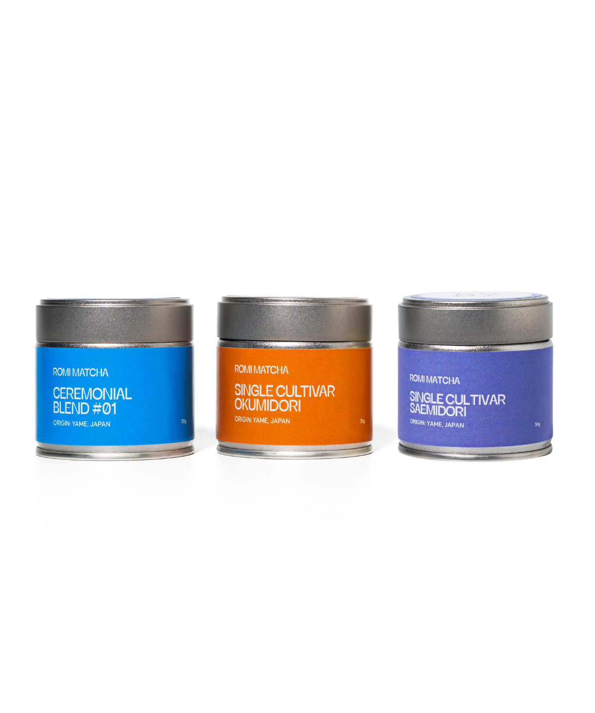 Three metal matcha tea canisters in blue, orange, and purple on a white background