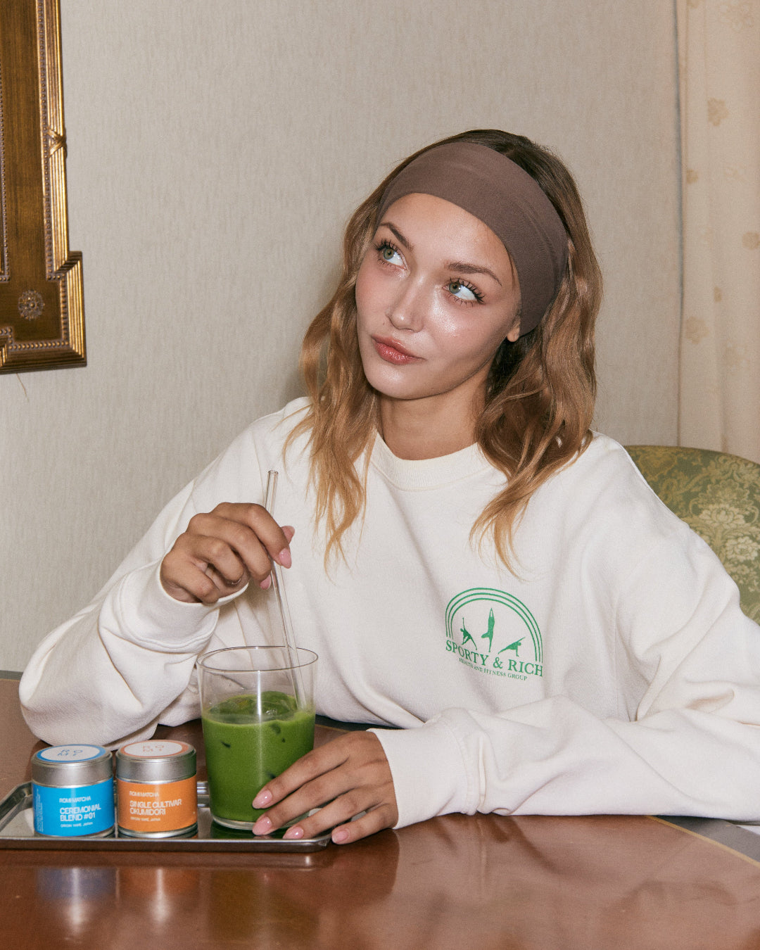 Woman sitting at a table with a green matcha latte and a white sweatshirt.