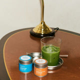 Two tins of matcha and a glass of green drink on a wooden table with a lamp in the background.