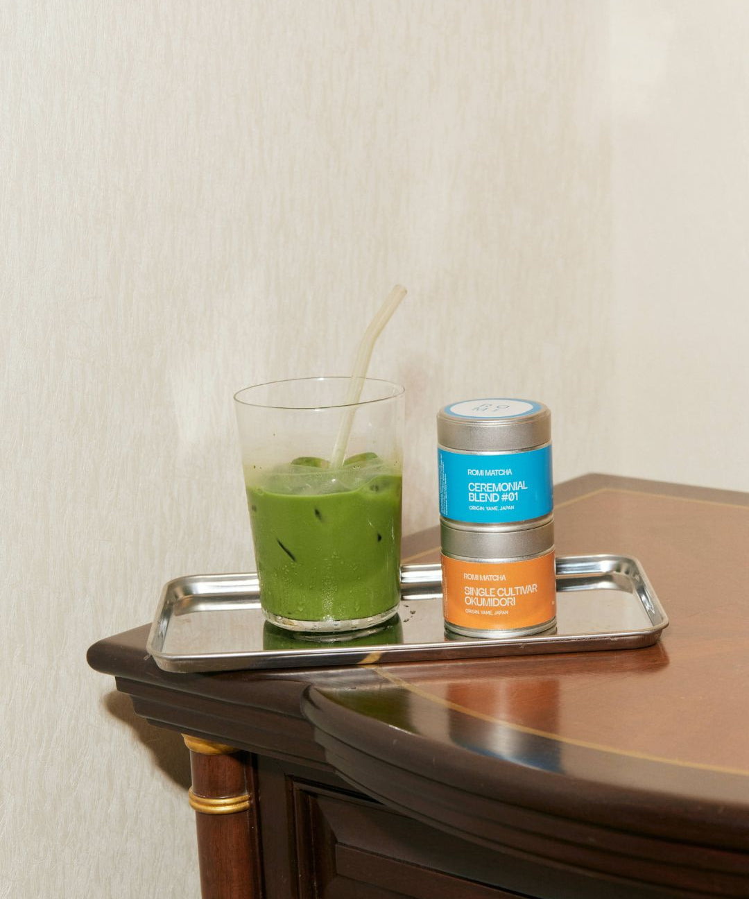 Green matcha drink in a glass with a straw on a tray next to matcha tins, on a wooden surface.