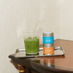 Green matcha drink in a glass with a straw on a tray next to matcha tins, on a wooden surface.