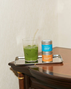 Green matcha drink in a glass with a straw on a tray next to matcha tins, on a wooden surface.